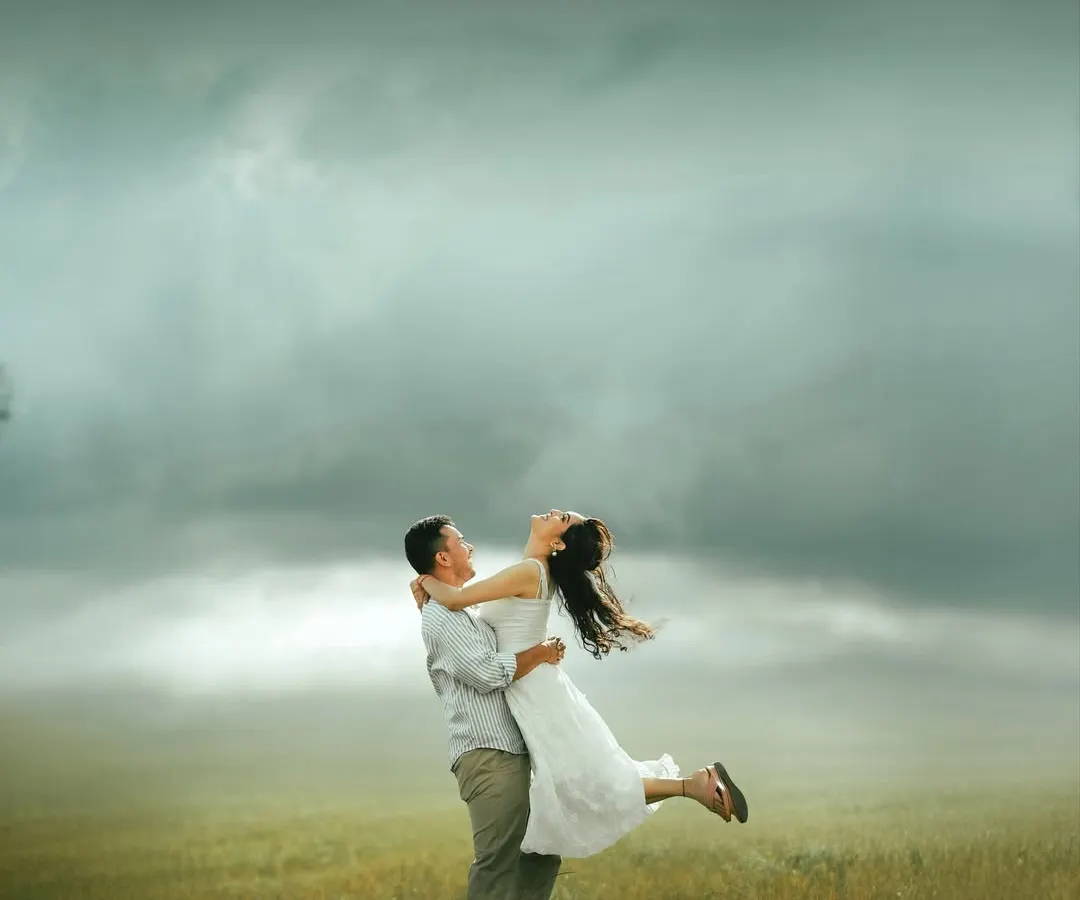 A joyful lift in an open field under a dramatic, cloudy sky. This image embodies the spontaneous and happy moments we aim to capture, using the vastness of nature to highlight the connection between two people. This is a perfect example of our 'dreamyframes' style.