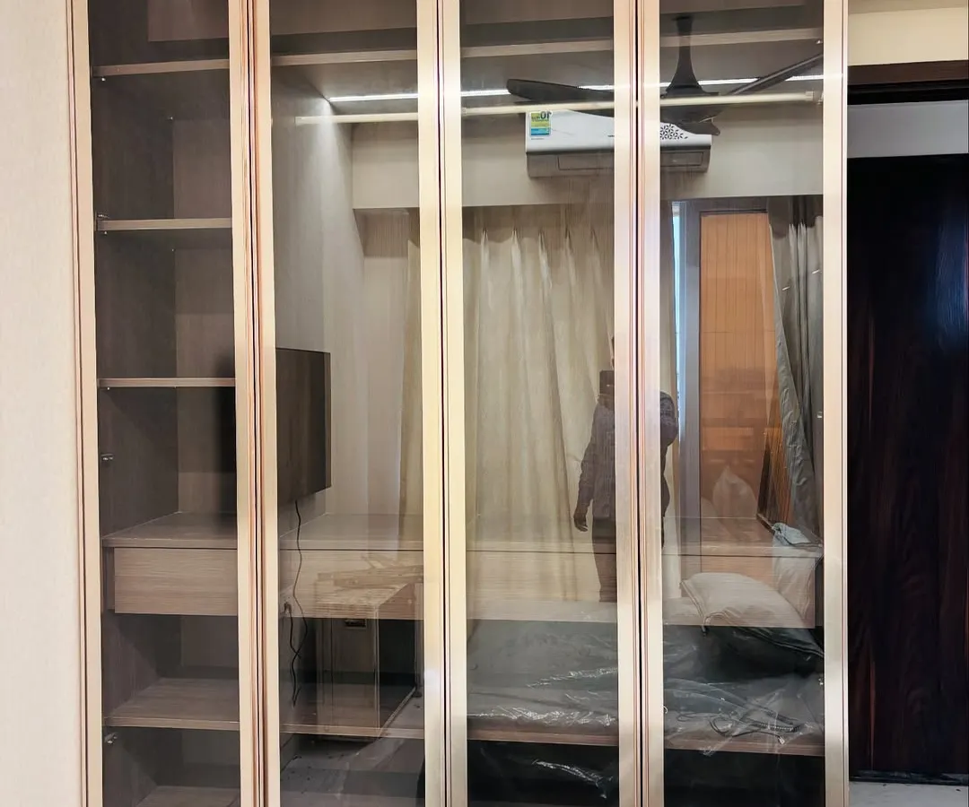 This elegant floor-to-ceiling wardrobe features tinted glass profile doors with a rich wood finish. The design offers a sophisticated way to display and organize your belongings, blending seamlessly into a modern bedroom aesthetic.