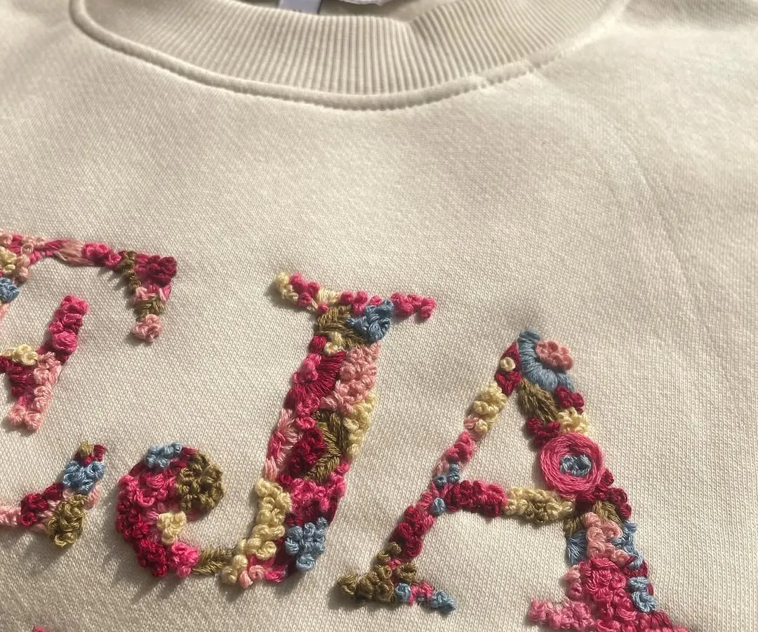 A close-up of the magic. I love filling initials with tiny, colorful flowers. Each petal and leaf is stitched by hand, making every floral letter a unique piece of art, like this 'TEJA' sweatshirt.