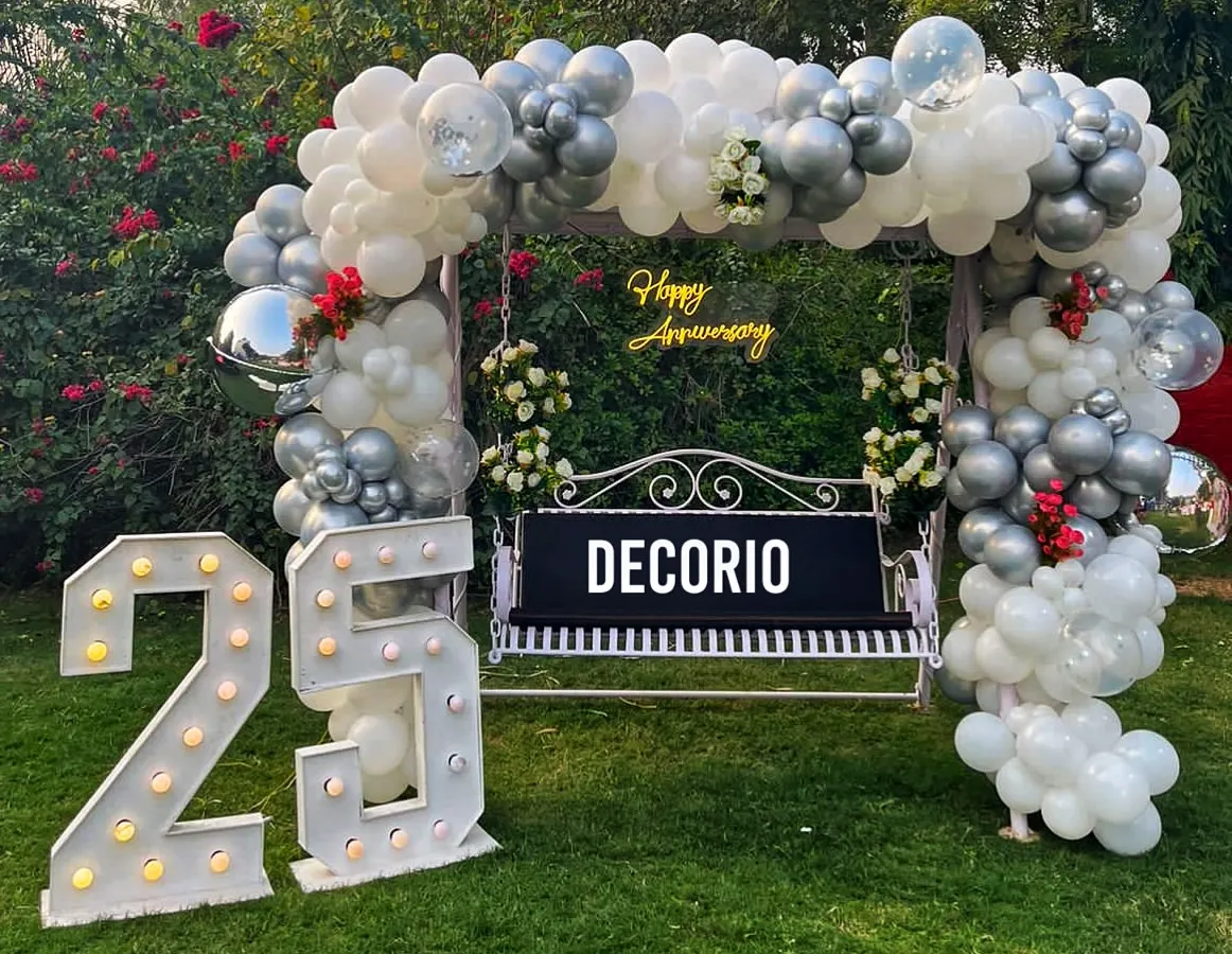For a milestone 25th anniversary, I created this elegant outdoor swing decoration. The setup features a lush silver and white balloon arch, a custom 'Happy Anniversary' neon sign, and large marquee numbers, making it a beautiful focal point for photos in a garden or lawn setting.