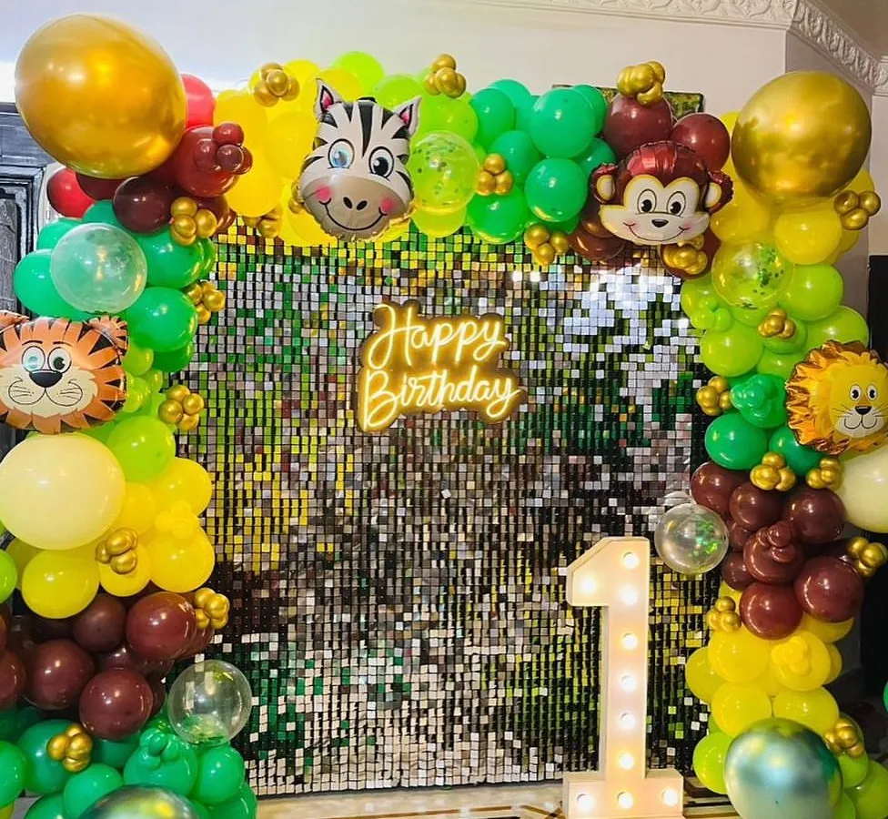 A vibrant jungle-themed first birthday party backdrop. This setup includes a shimmering sequin wall, a custom neon sign, marquee number, and a lush balloon garland with fun animal characters.