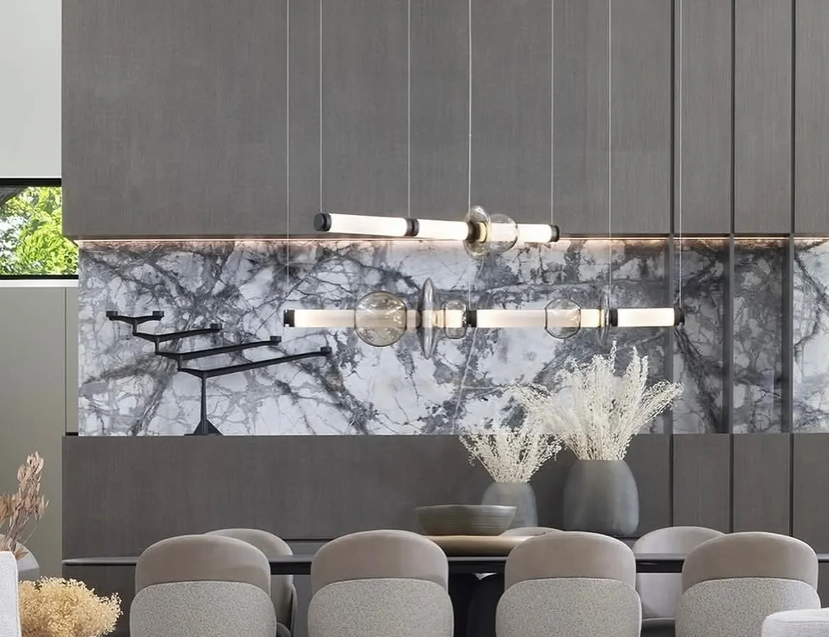 The Luna series by Gabriel Scott demonstrates how linear lighting can become the centerpiece of a grand dining space. Its modular design, featuring blown glass and metallic finishes, is suspended against a dramatic marble wall, creating a statement of sophisticated, contemporary luxury.