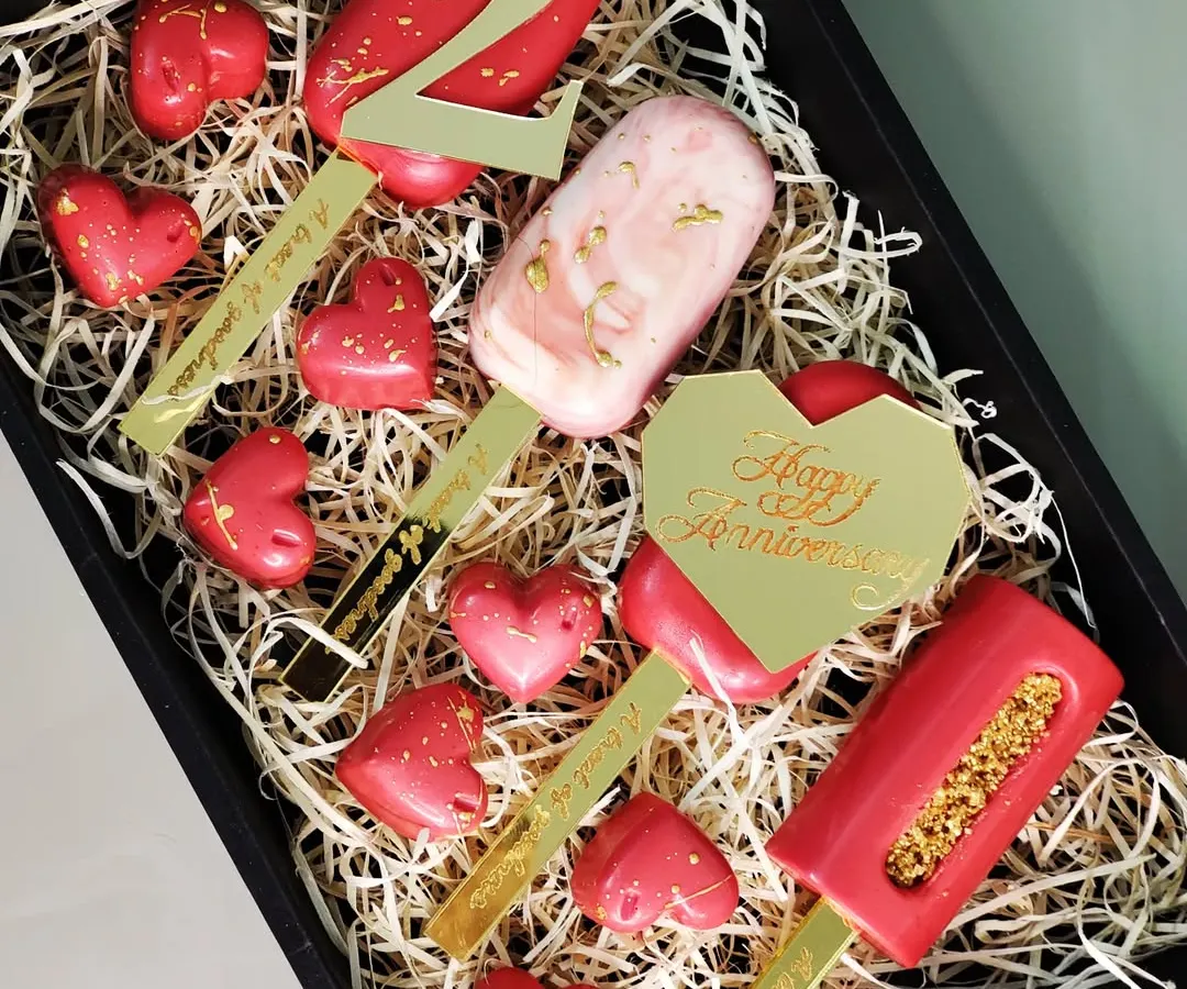 A personalized anniversary gift box designed to celebrate a special milestone. This set features custom-engraved cakesicles, including a number '2' and a 'Happy Anniversary' plaque, paired with heart-shaped chocolates.