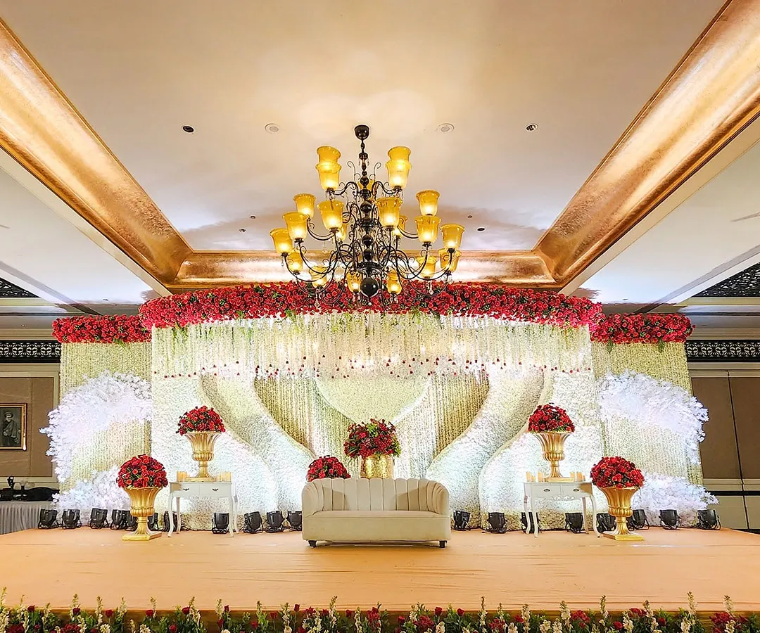 We specialize in creating opulent reception stages that serve as the magnificent centerpiece of your celebration. This design features a grand floral canopy of red and white roses with cascading crystal elements.