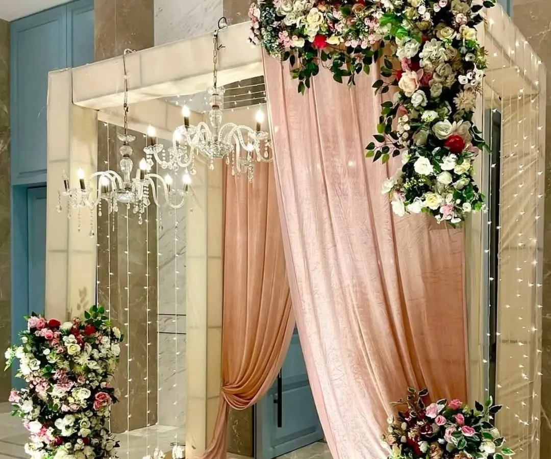An elegant and grand entrance designed for a wedding or reception. The decor features soft pink draping, cascading floral arrangements with roses and greenery, and sparkling chandeliers that create a luxurious and welcoming ambiance for your guests.