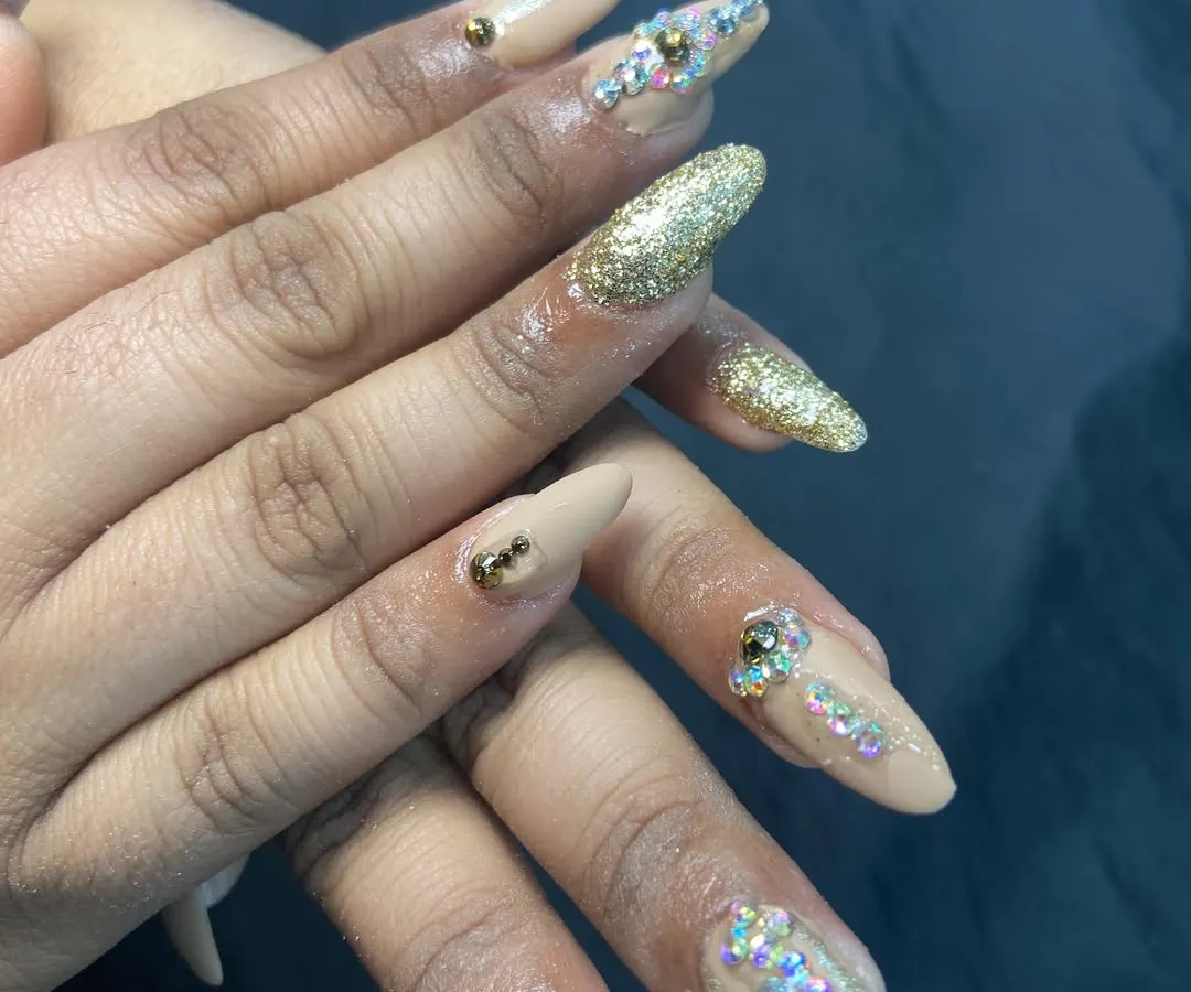 For when you want to make a statement. These long, almond-shaped nail extensions feature a mix of nude polish, dazzling gold glitter, and carefully placed rhinestones for a truly luxurious and festive feel.