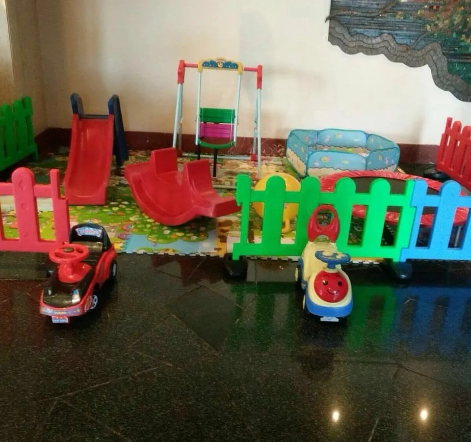 This is an example of an indoor play area setup on a hard floor. It features a slide, swing, rocker, ride-on cars, and a foldable ball pit, all enclosed by a colorful fence.