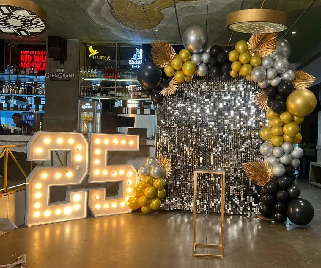 For a 25th anniversary party, I created this glamorous photo corner. It includes a shimmering silver sequin backdrop, large light-up '25' marquee numbers, and a custom balloon arch in a sophisticated black, gold, and silver color scheme.