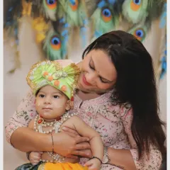 A loving portrait of a mother holding her baby, who is dressed in a Krishna costume against a backdrop of peacock feathers.