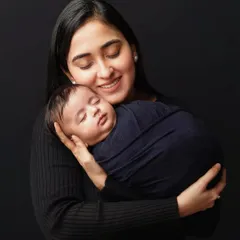 A mother smiles with her eyes closed, holding her swaddled newborn in her arms. The baby is wrapped in a dark blue fabric that complements the simple black outfits and background.