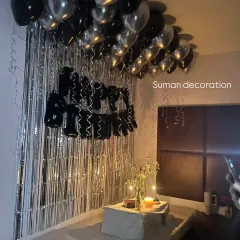 A romantic birthday surprise in a bedroom using black and silver balloons. The setup includes a silver tinsel curtain and a small table for a cake, creating an intimate celebration space.