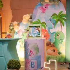 A close-up of the dessert table area for Avyaan's dino party. The setup includes a custom backdrop with cartoon dinosaurs and themed pedestals for the cake and treats.