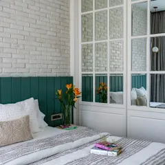 This cozy bedroom from the 'Urban Oasis' project features a white brick accent wall and a deep green channel-tufted headboard. The mirrored wardrobe doors add depth and reflect light, making the room feel larger.
