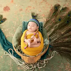 A creative and cultural newborn portrait of a baby dressed as Little Krishna. The baby rests in a basket designed to look like a peacock, complete with a full feather tail, holding a tiny flute.