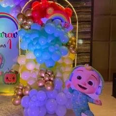 A vibrant Cocomelon theme for a first birthday party. This setup includes a custom arched backdrop, a rainbow-colored balloon arrangement, and a life-sized cutout of the character JJ.