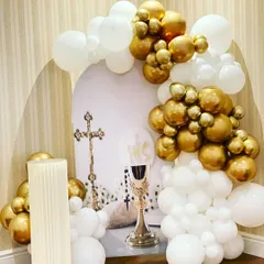 An elegant First Holy Communion setup in white and gold. The decor includes a balloon garland framing a backdrop with religious symbols like the cross and chalice.