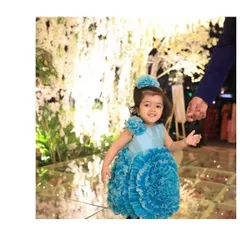 A vision in blue. This custom flower ball dress features a skirt made of countless handcrafted organza ruffles, creating a stunning, voluminous look. It’s paired with a matching floral headband for a picture-perfect first birthday outfit.