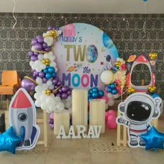A 'Two the Moon' space themed second birthday party. This decoration features a circular backdrop, a purple and blue balloon garland, and astronaut and rocket cutouts.