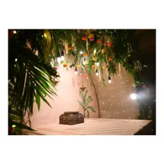 This image showcases the elegant Ganpati mandap we designed, featuring a custom-carved wooden seat awaiting the idol. The space is framed by an arch of fresh greenery and flowers, with hanging bulbs and fairy lights creating a warm, inviting glow against the hand-painted wall.