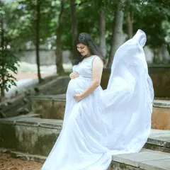 An expectant mother looking serene in a flowing white gown during an outdoor maternity session. The natural park setting and the gentle toss of the fabric create a timeless and angelic portrait.