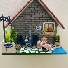 This custom diorama for 'Nik & Mili' captures a romantic scene. It features handmade clay miniatures of the couple, a tiny table set for two, and a welcoming fence, all against a detailed brick-textured house.