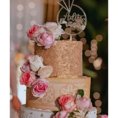 A stunning three-tiered engagement cake, adorned with gold glitter, intricate lace patterns, and fresh pink roses, topped with a custom 'Aditi & Pranav' ring topper.