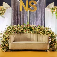 A close-up of a modern Sangeet stage with a personalized "NS" monogram, a shimmering crystal curtain, and a base of pastel-colored flowers.