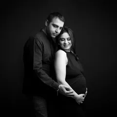 A classic and elegant black and white portrait. The couple embraces tenderly, their poses conveying a sense of love, protection, and shared anticipation.