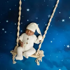 A dreamy portrait of a newborn in a white knit outfit, appearing to sleep on a macrame swing against a starry night sky backdrop.