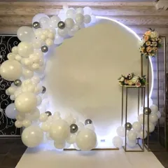 A modern and chic baptism decoration with a backlit circular backdrop. The setup is framed by a white and silver balloon arch and accented with floral bouquets.