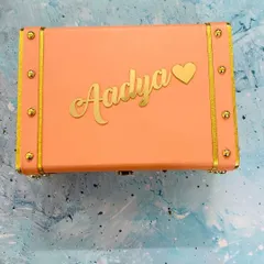 A lovely peach-colored trunk box, customized with a name in elegant gold script and a matching heart charm. It's a beautiful and personal way to store your valuables.