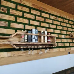 A detailed close-up of the bottle-shaped bar shelf. This view highlights the craftsmanship, the integrated stemware rack for wine glasses, and the textured background combining wood blocks and green turf.