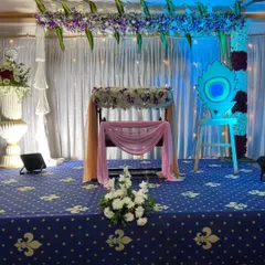 This naming ceremony stage features a traditional cradle, or thottilu, decorated with flowers and drapes. The backdrop includes a peacock cutout and an overhead floral arrangement with hanging leaves.