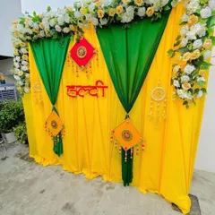 A simple yet elegant Haldi backdrop featuring a combination of yellow and green drapes. The top is lined with a garland of artificial white and yellow roses, adding a touch of floral beauty to the traditional setup.