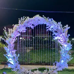 A circular photo arch decorated with white cherry blossoms and illuminated with soft purple light. The hanging crystal strings in the center add a touch of sparkle, making it a perfect selfie spot.