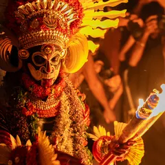 A close-up of the Karim Gullikan Theyyam, holding a torch that illuminates the intricate details of his mask and costume. The firelight reveals the textures and colors that bring the deity to life.