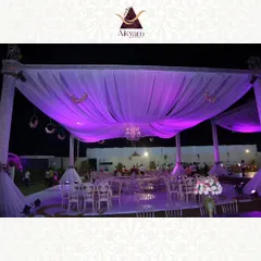An expansive outdoor reception setup under a canopy of draped white fabric, illuminated by dramatic purple uplighting and a central crystal chandelier. The scene is set for a grand and elegant evening celebration.