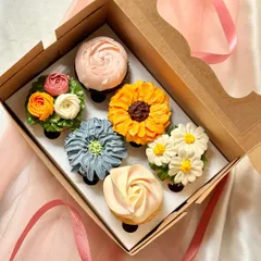 A gift box of six cupcakes decorated with intricate, multi-toned buttercream flowers. The collection displays a variety of floral piping techniques, including a sunflower, daisies, a rose, and other blossoms, perfect for elegant events like birthdays or anniversaries.