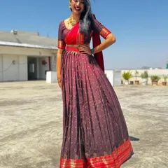 This half-saree features a deep maroon skirt with a traditional gold and red border, paired with a contrasting red blouse and dupatta for a vibrant, celebratory look.