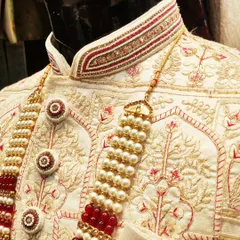 The difference is in the details. A close-up of a bespoke sherwani collar, showcasing intricate hand embroidery with red thread, pearls, and zardosi work on an ivory base, complete with custom-designed buttons.