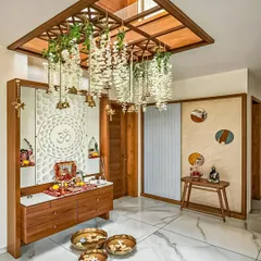 This elegant pooja space features a mandir with a white 'Om' jaali panel and a wooden cabinet. The ceiling is adorned with a wooden lattice from which brass bells and floral hangings are suspended.