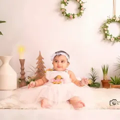 A beautiful portrait using a minimalist, boho-inspired setup. The baby is dressed in a lovely white dress, surrounded by potted plants and natural textures for a clean, modern look.