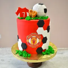 A cake for a true football fan. This design is dedicated to Manchester United, featuring the team's logo, a jersey, and soccer balls, all made from edible fondant.