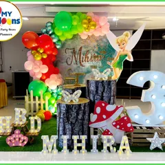 A whimsical fairy-themed third birthday party. The decor includes a Tinkerbell cutout, a forest backdrop, log-style plinths, and a large light-up number '3', creating an enchanted woodland scene.