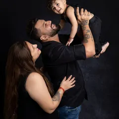 A dynamic and joyful family portrait. The father lifts his child high in the air, creating a moment filled with laughter and energy, all captured against a classic dark backdrop.