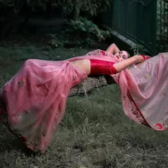 An artistic and dramatic pose, with the model lying on a fallen tree log in a lush green setting. The pink saree drapes beautifully, creating a stunning contrast with the natural surroundings.