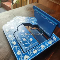 A close up of a Jharokha mirror with its shutter opened. The white floral painting on the blue base is a classic design that adds elegance to any wall.