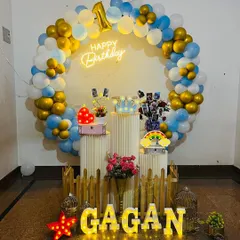 A beautiful ring backdrop for a first birthday celebration. The balloon ring in blue, white, and gold chrome frames a display of photos and props on plinths, with a 'Happy Birthday' neon sign and light-up name.
