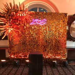 A glamorous photo booth for a sangeet or cocktail night. The backdrop is made of shimmering gold sequins, complemented by a personalized neon name sign and dramatic floral accents.