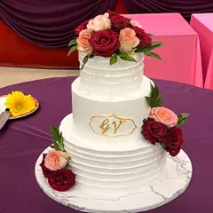 A classic three-tier white wedding cake with a simple, elegant ridged texture. It's decorated with a cascade of fresh red and peach roses and features a custom gold monogram, proving that true beauty lies in simplicity.
